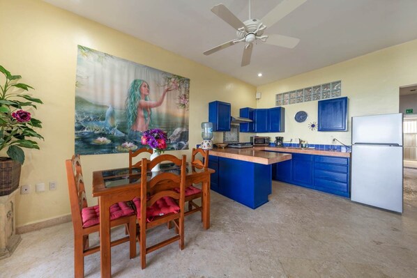 Una cocina con armarios azules, una mesa de comedor con sillas, un ventilador de techo y un cuadro en la pared.
