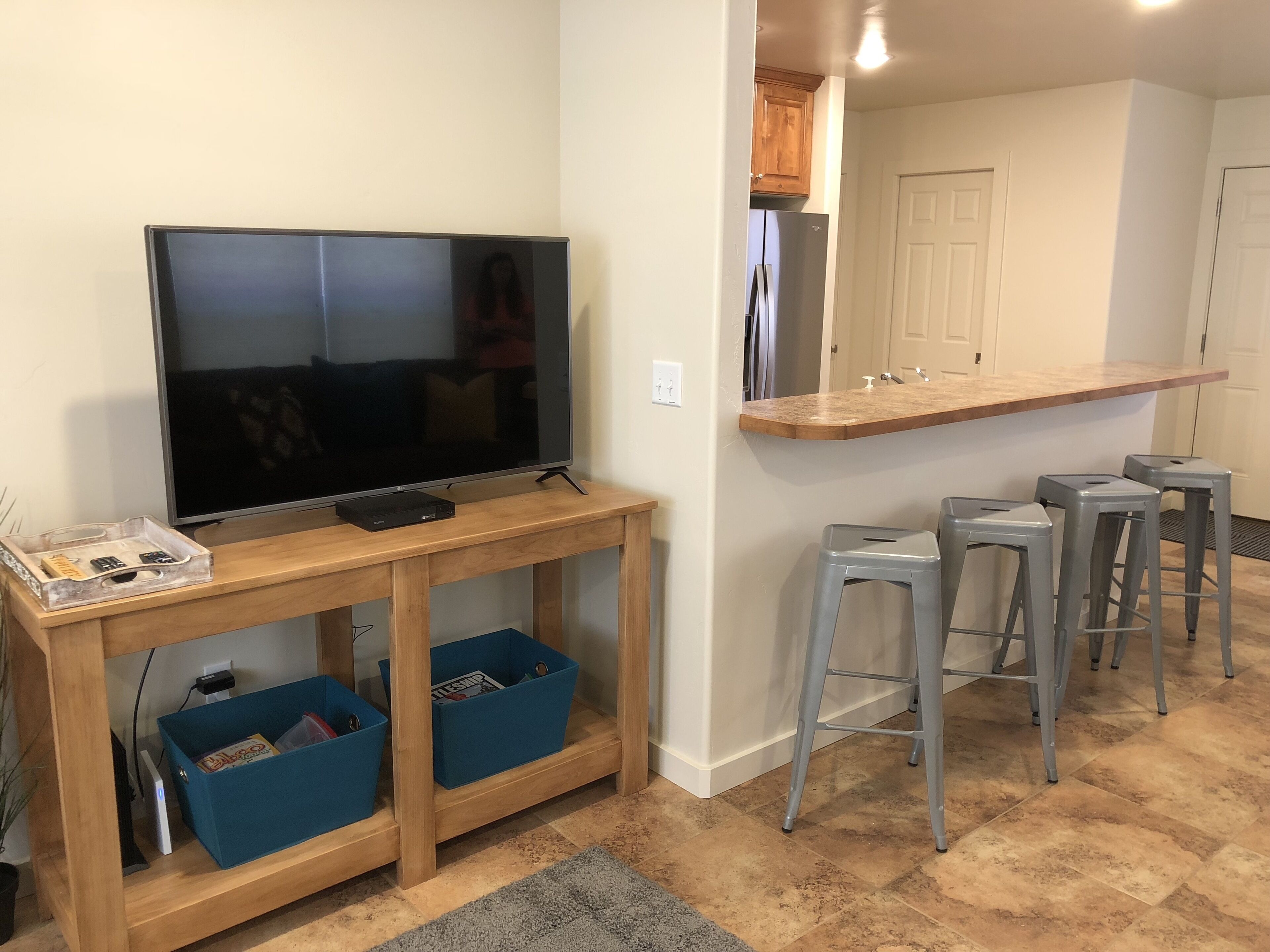 A living room with a TV on a wooden stand, blue storage bins, and a kitchen area with a refrigerator and bar stools.