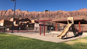 Ein Spielplatz mit Rutsche und Klettergerüsten, umgeben von Gebäuden und einer Bergkulisse.