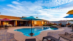 A swimming pool with lounge chairs and umbrellas, surrounded by a brick patio and a building with outdoor seating. - Tequila Sunrise, Large Pool Home (Lake Havasu City)