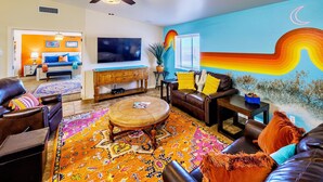 A living room with a large TV, a ceiling fan, a colorful rug, and a wall mural. - Tequila Sunrise, Large Pool Home (Lake Havasu City)