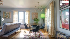 A modern living room with a sofa, dining table, chairs, a green floor lamp, and a large window with curtains.