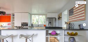 Una cocina moderna con armarios blancos, una isla central con taburetes de bar y objetos decorativos en estantes.