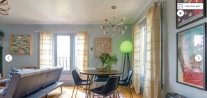 Una sala moderna con un sofá, una mesa de comedor, sillas, una lámpara de pie verde y un ventanal con cortinas.