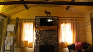 A log cabin interior with a stone fireplace, a mounted television, and a wooden ceiling.