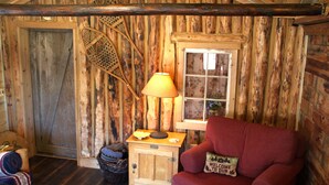A rustic cabin interior with a wooden door, a window with floral curtains, a red sofa with a 'Welcome' pillow, a lamp on a wooden table, and a basket with blankets.