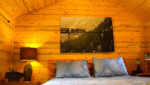 A wooden cabin room with a bed, two patterned pillows, a nightstand with a lamp, and a painting of a bridge over a forest.