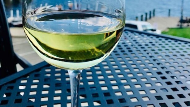 A glass of white wine on a metal table with a view of a body of water and a bridge in the background.