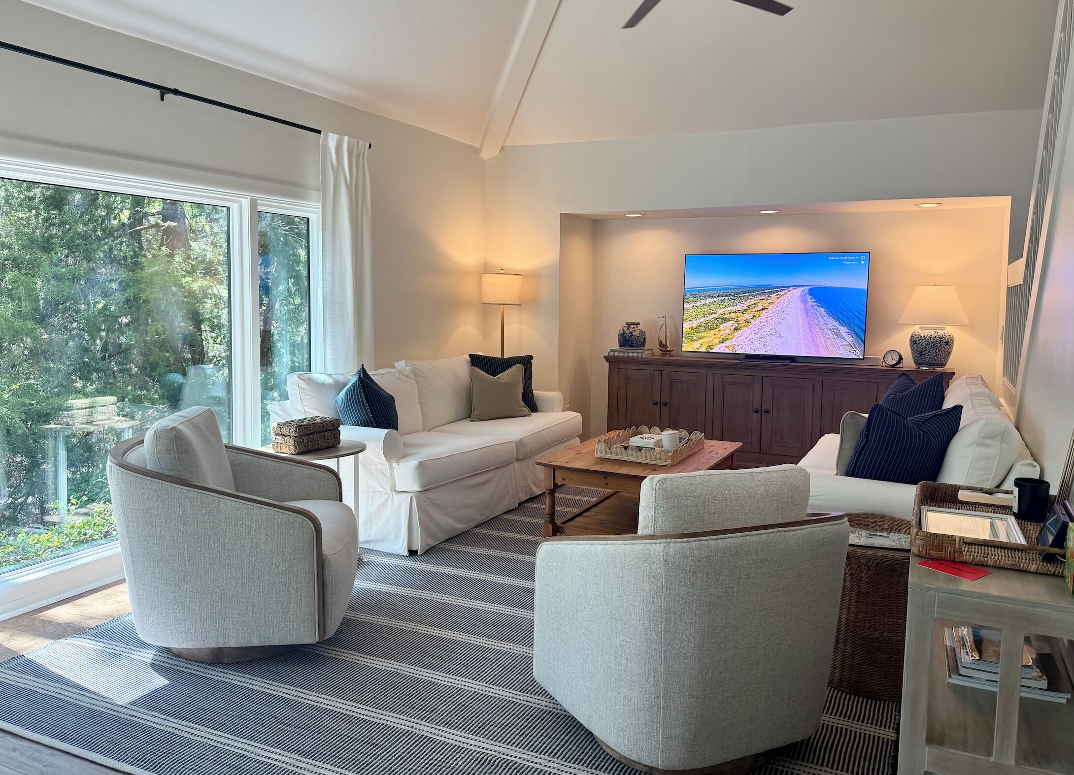Ein modernes Wohnzimmer mit einer Couch, Sesseln, einem Couchtisch, einem Flachbildfernseher und Blick auf einen Strand durch große Fenster.