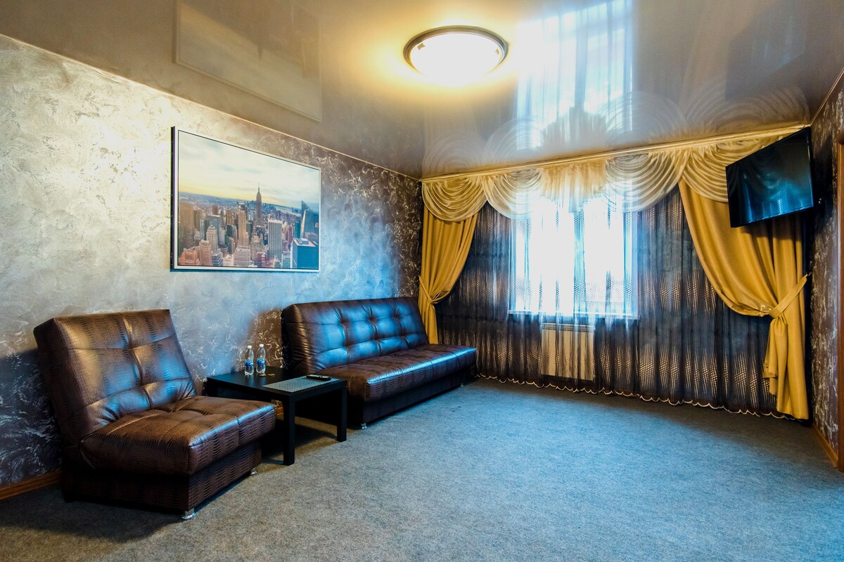 A room with a brown leather sofa, a brown leather armchair, a small table with a remote control, and a framed cityscape picture on the wall.