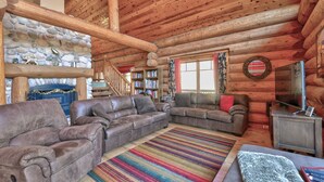 A cozy log cabin interior with leather sofas, a stone fireplace, a bookshelf, and a television.