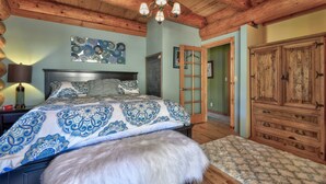 A bedroom with a wooden ceiling, a bed with patterned bedding, a white furry ottoman, and a wooden wardrobe.