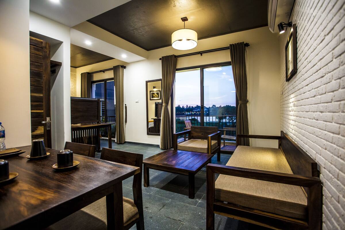 A modern hotel room with a wooden dining table, two wooden chairs, a sofa, and a large window with curtains.