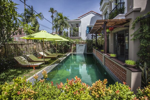 A swimming pool with lounge chairs and umbrellas, surrounded by lush greenery and a white building with a balcony. - Hoi An Silk River Villa (Da Nang)