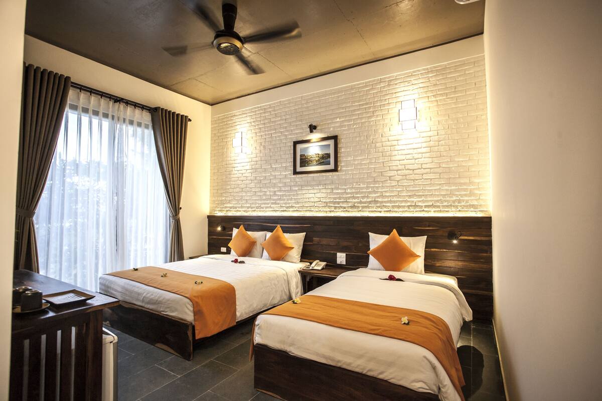 A hotel room with two beds, a desk, a ceiling fan, and a brick-patterned wall with a framed picture.