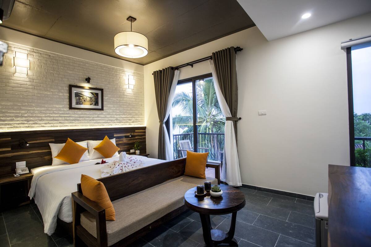 A hotel room with a large bed, a bedside table, a round table with a plant, and a window with curtains.