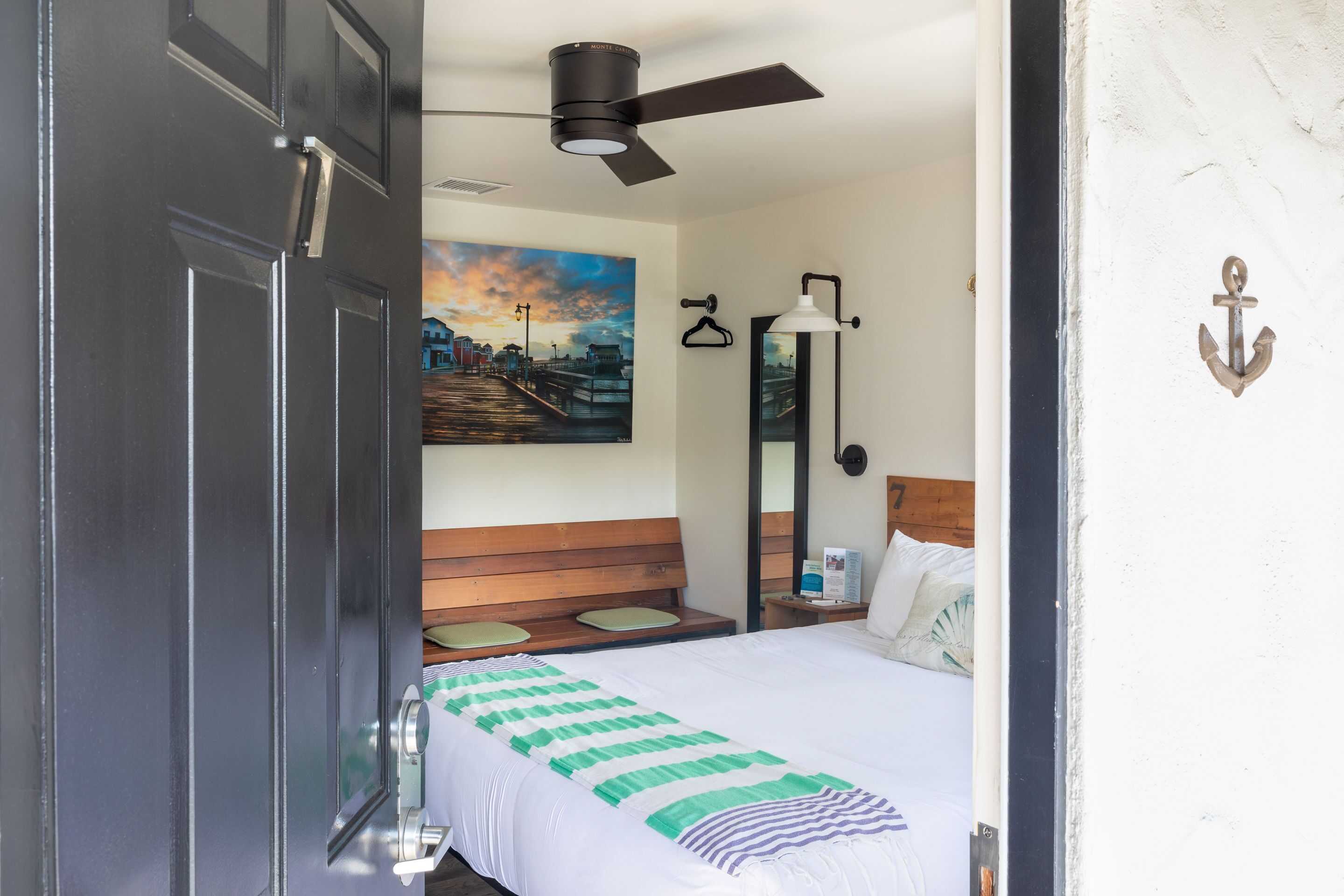Ein Hotelzimmer mit einem Bett, einem Deckenventilator, einem Sonnenuntergangsbild, einem hölzernen Kopfteil, einem Nachttisch und einem Spiegel.