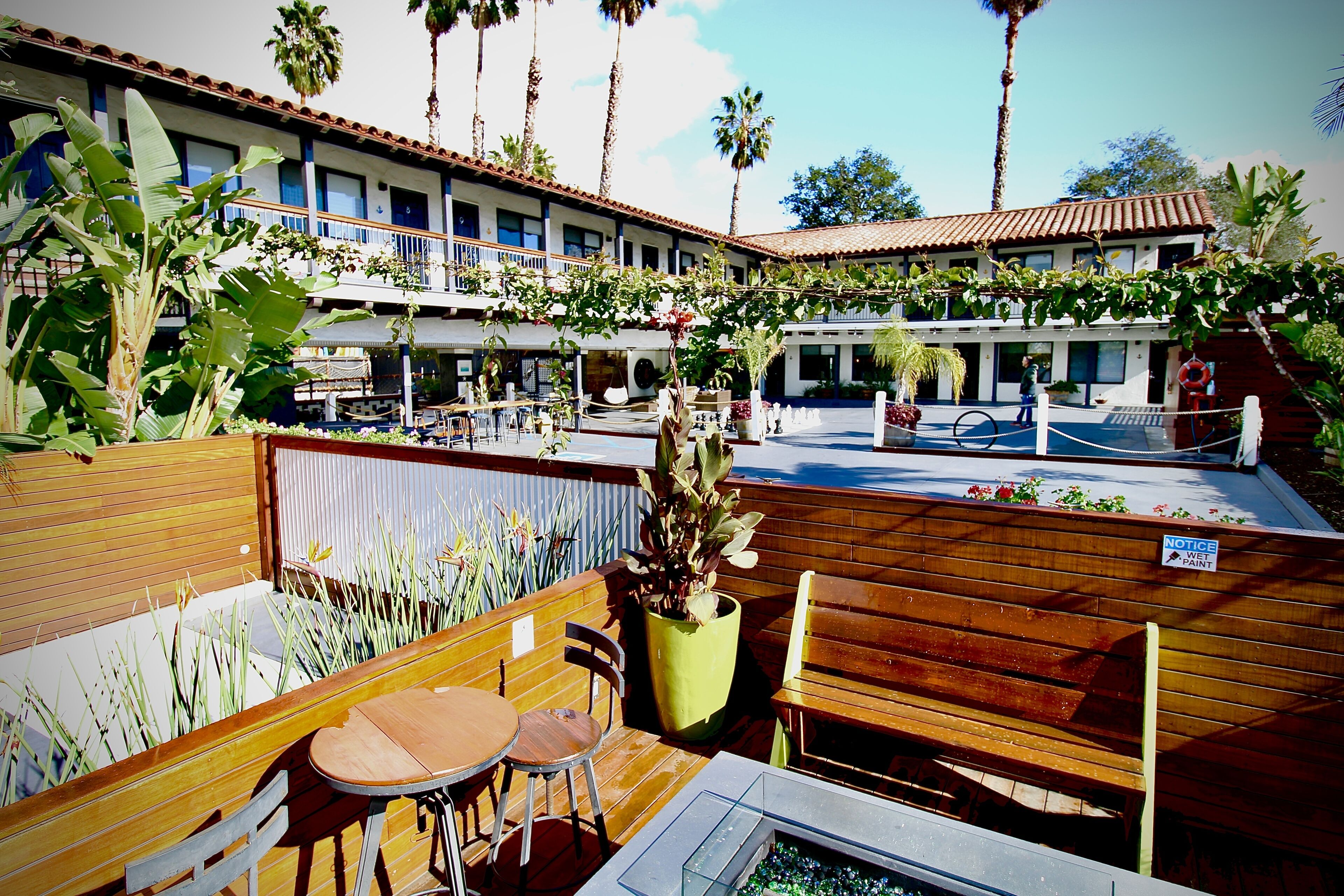 Un patio al aire libre con mobiliario de madera, una piscina y un edificio con múltiples ventanas y balcones.