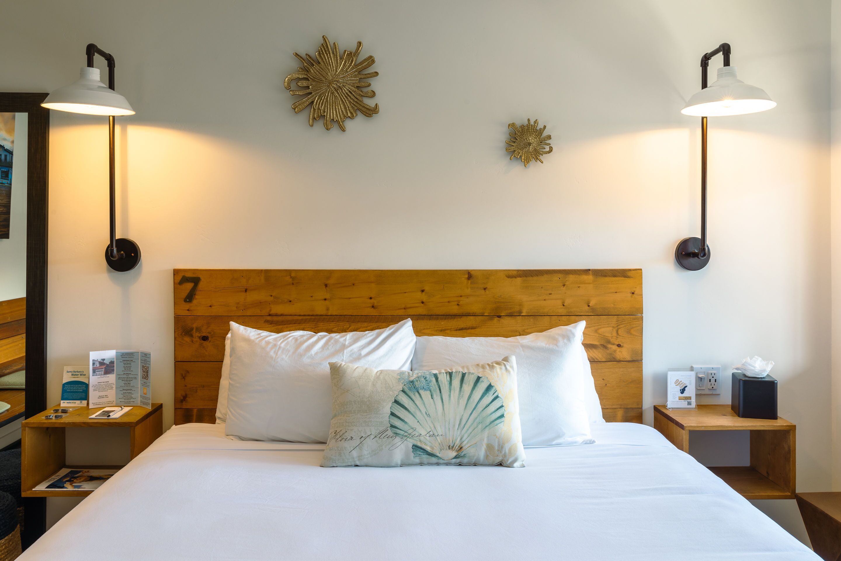 A hotel room with a wooden headboard, two white pillows, a decorative pillow with a fan design, two bedside tables with lamps, and wall art.