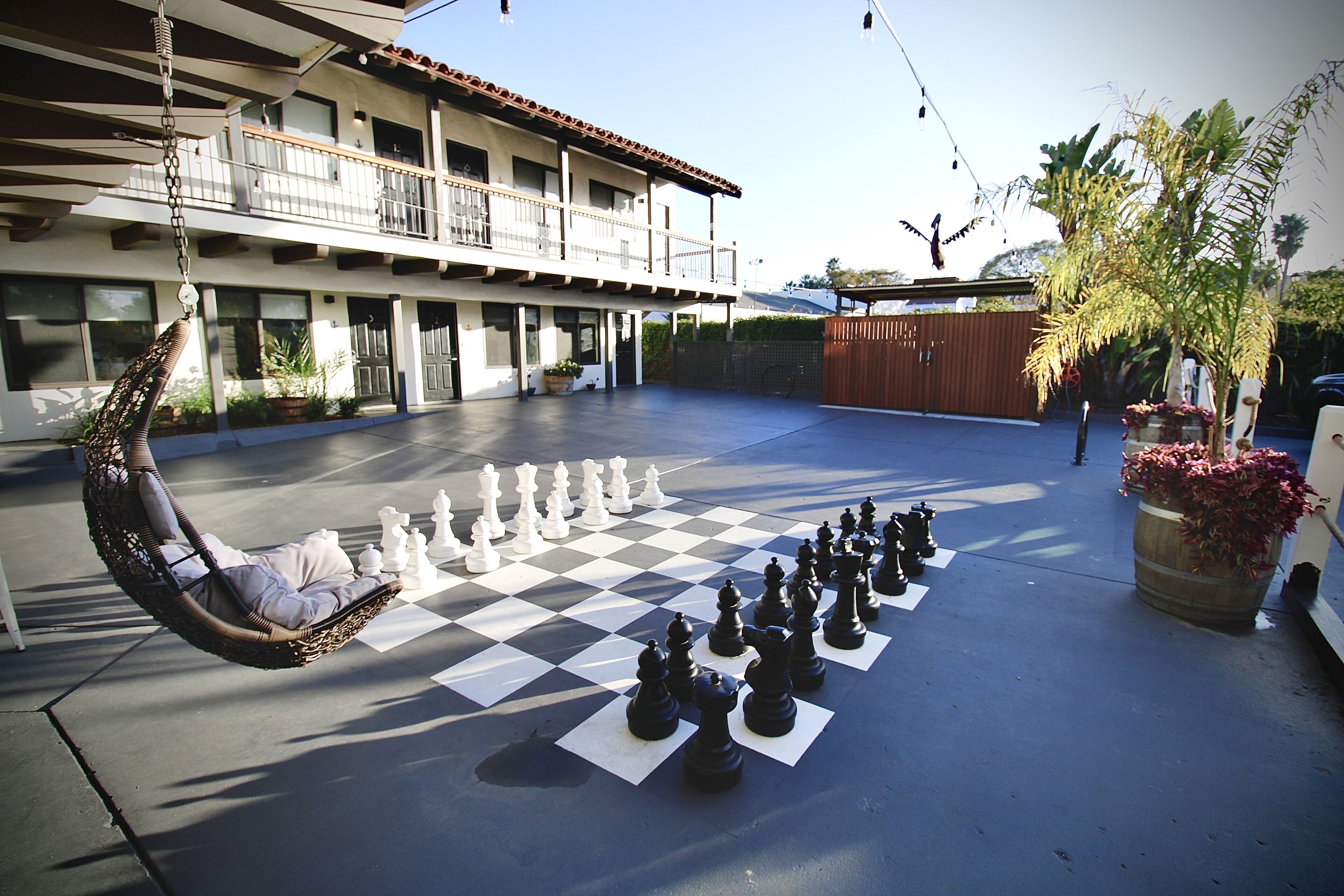 A large outdoor chessboard set up on a courtyard with a hanging chair and potted plants.