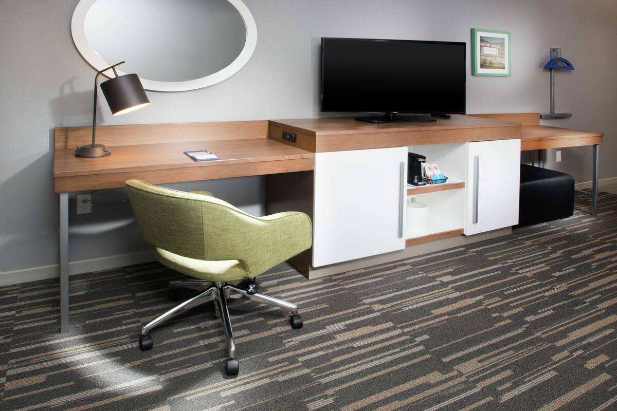 Habitación de hotel moderna con un escritorio de madera, una silla de oficina verde, un televisor de pantalla plana, un espejo redondo y una mesita de noche pequeña.