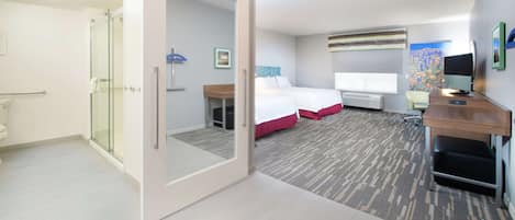 Habitación de hotel con cabina de ducha de vidrio, cama con ropa de cama roja, escritorio con computadora y un ventanal grande.