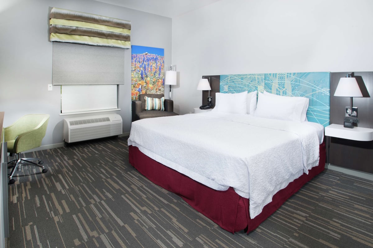 Habitación de hotel con una cama grande, un escritorio con una silla, una lámpara de escritorio, un aire acondicionado montado en la pared y un cuadro.