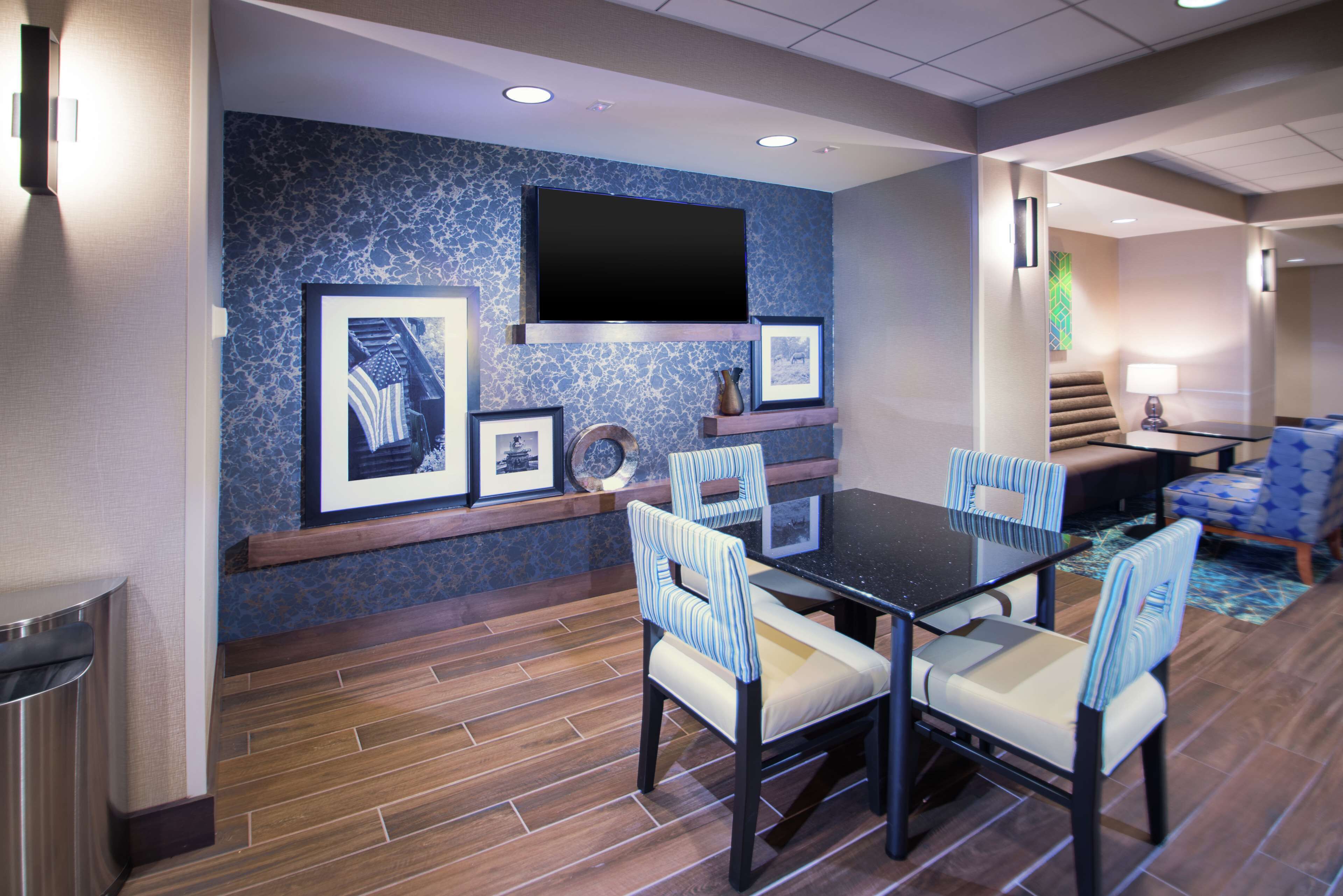 A hotel room with a dining area featuring a black table and striped chairs, a mounted flat-screen TV, framed artwork, and a patterned wall.