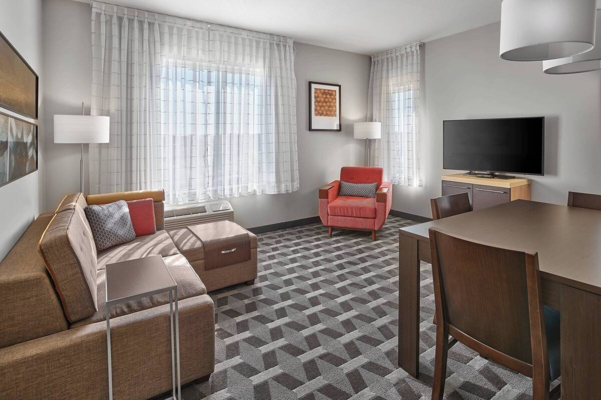 A modern hotel room with a sofa, armchair, dining table, and TV.