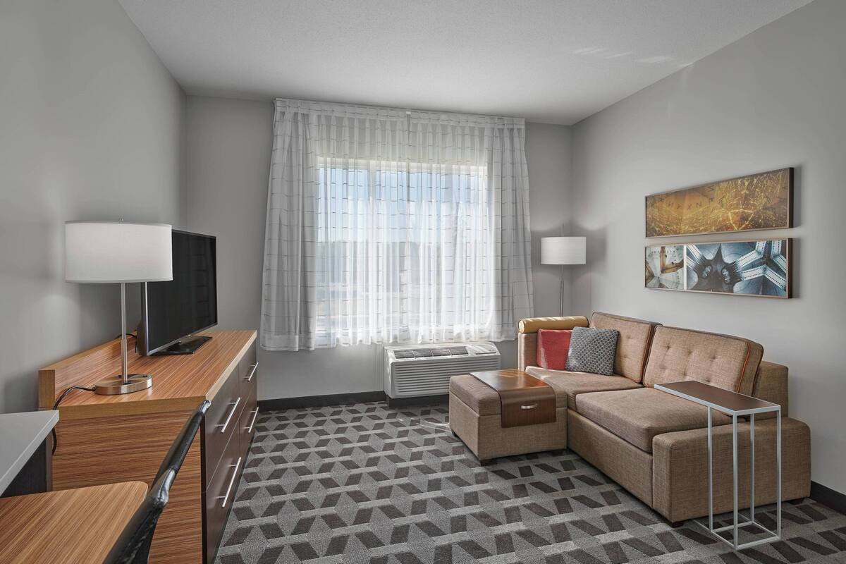 A modern hotel room with a sofa, a TV, a desk, and a patterned rug.