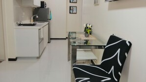 Une cuisine compacte avec des meubles blancs, un petit plan de travail et une cuisinière encastrée. On y trouve une table en verre avec une chaise, une plante en pot et un tableau encadré accroché au mur.