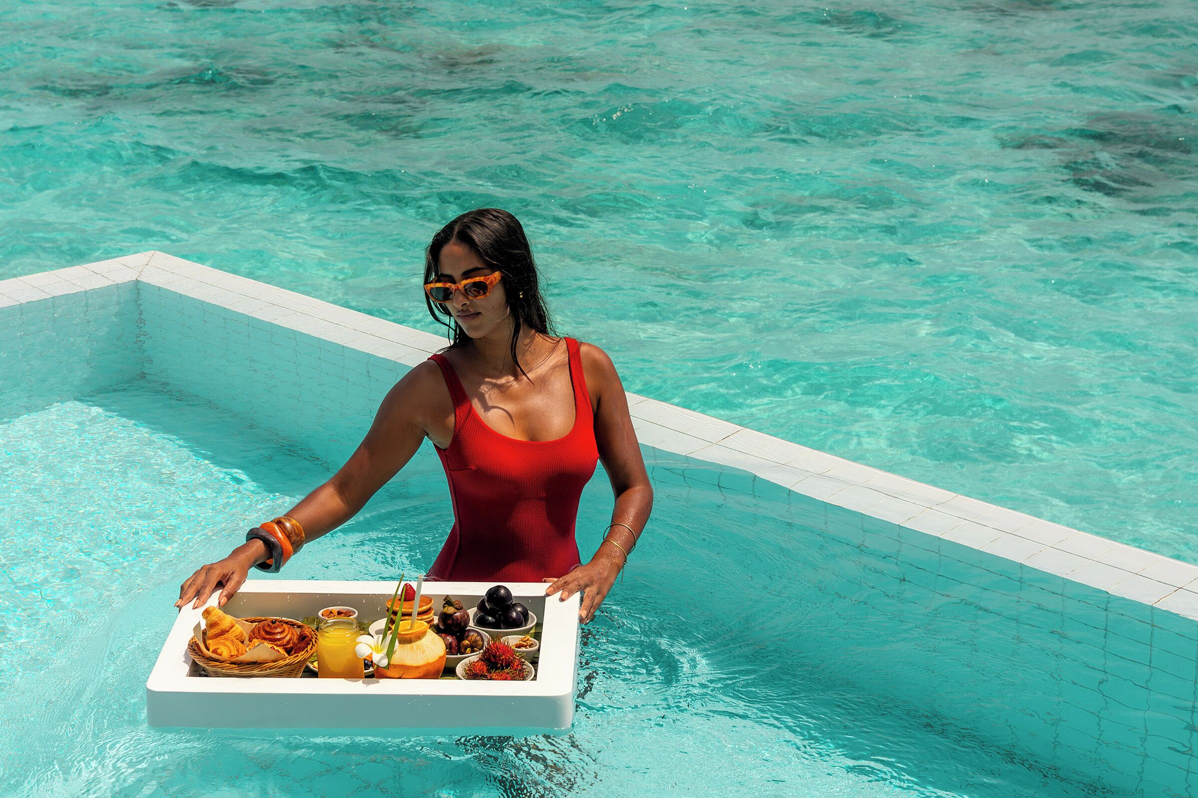A person in a red swimsuit serving a tray with food and drinks by a pool.