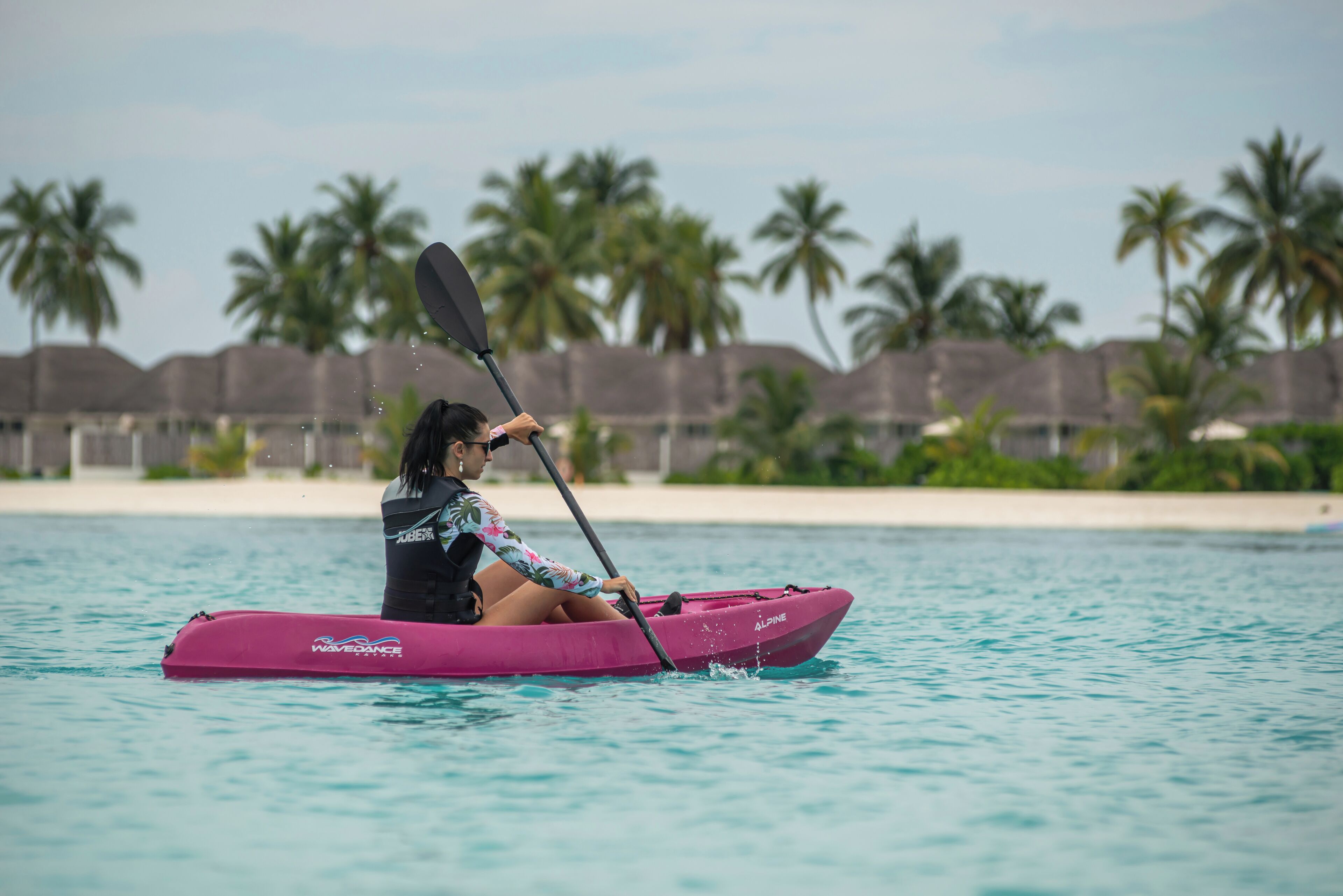 A person in a pink kayak on a calm body of water with palm trees and a beach in the background.