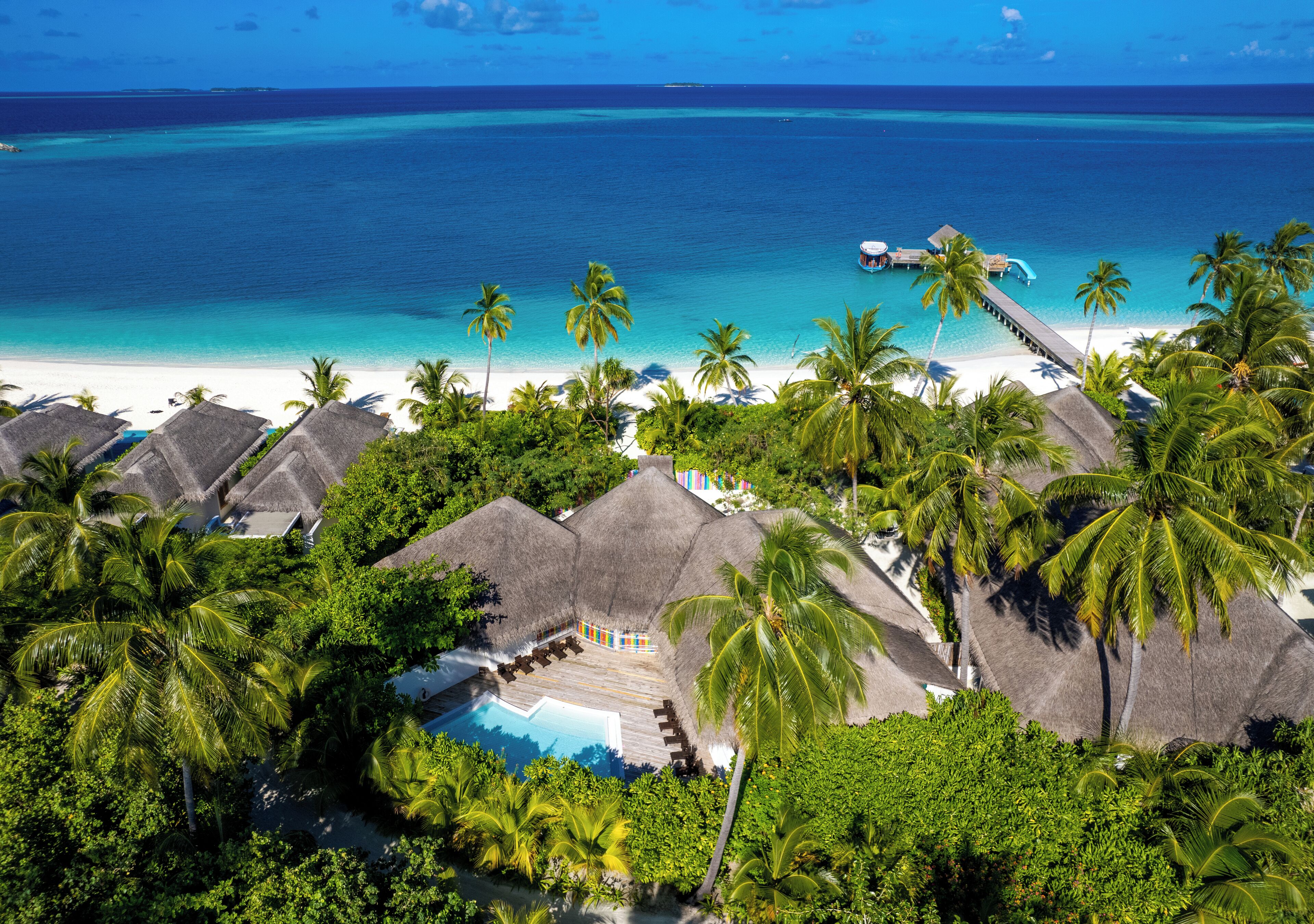 Vista aérea de un resort tropical con bungalós sobre el agua, una piscina y una playa.