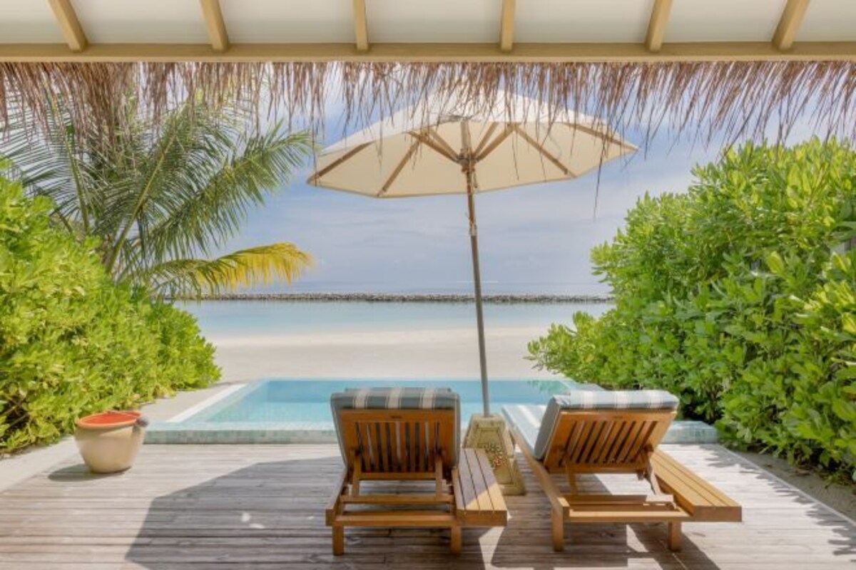 A wooden deck with two lounge chairs and a large umbrella, overlooking a pool and beach.