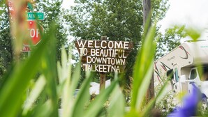 Un cartel de bienvenida a Talkeetna centro, con una caravana blanca estacionada cerca.