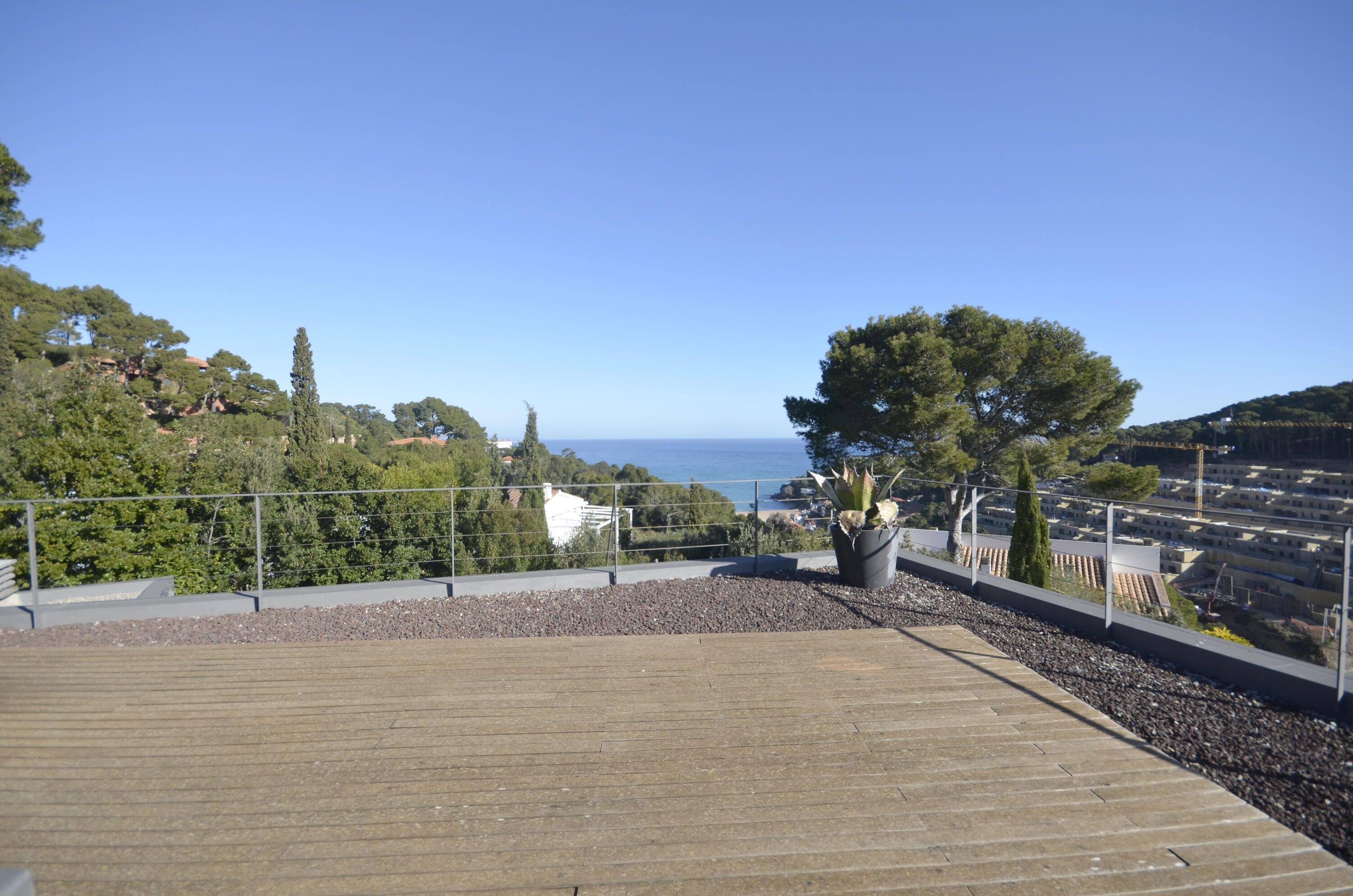Terraza de madera con barandilla, una planta en maceta y vista al mar y las colinas.