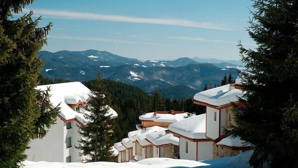 Ein schneebedecktes Bergresort mit Chaletgebäuden, freier Aussicht auf die umliegenden Berge und einem bewaldeten Hintergrund.
