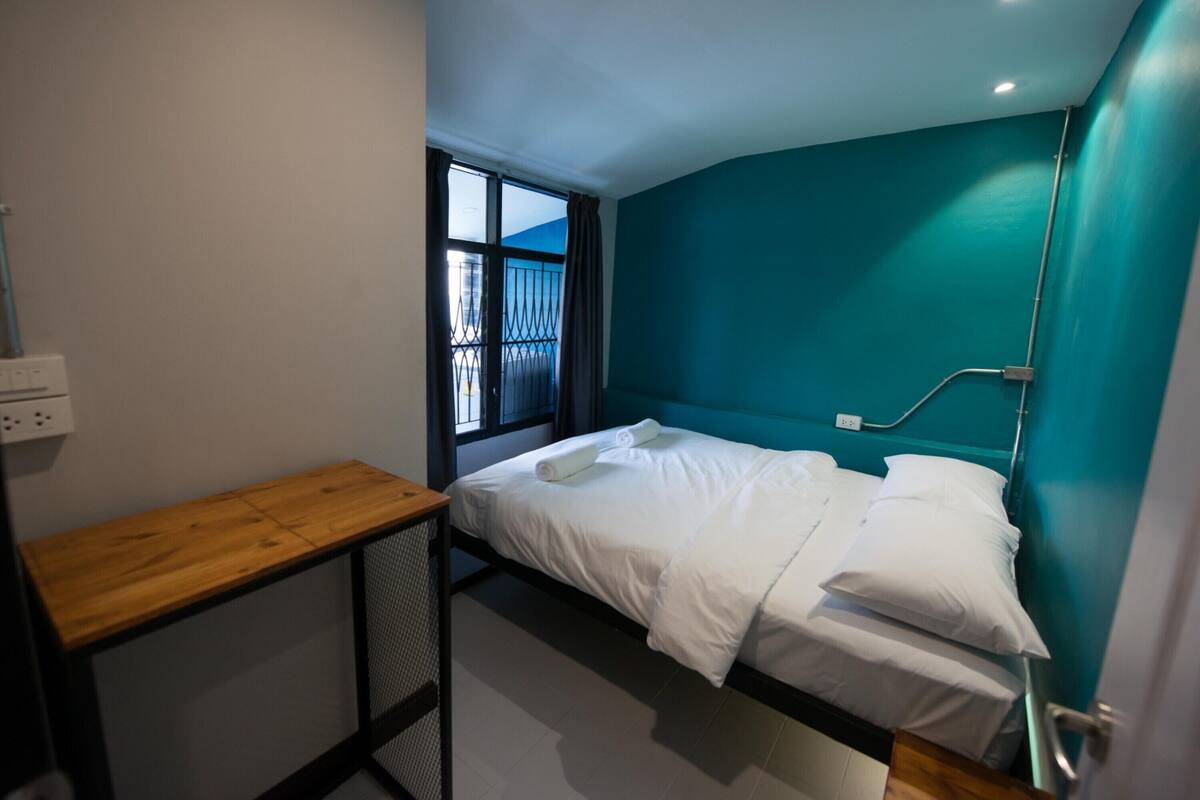 Ein kleiner, moderner Raum mit einem Einzelbett, einem Holztisch und einem Fenster mit Blick nach außen.