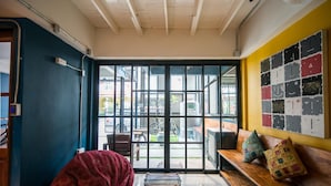 Una habitación con un ventanal grande, un sillón de girable tipo saco de frijoles rojo, un banco de madera con cojines de colores, y una pared con una cuadrícula de imágenes.