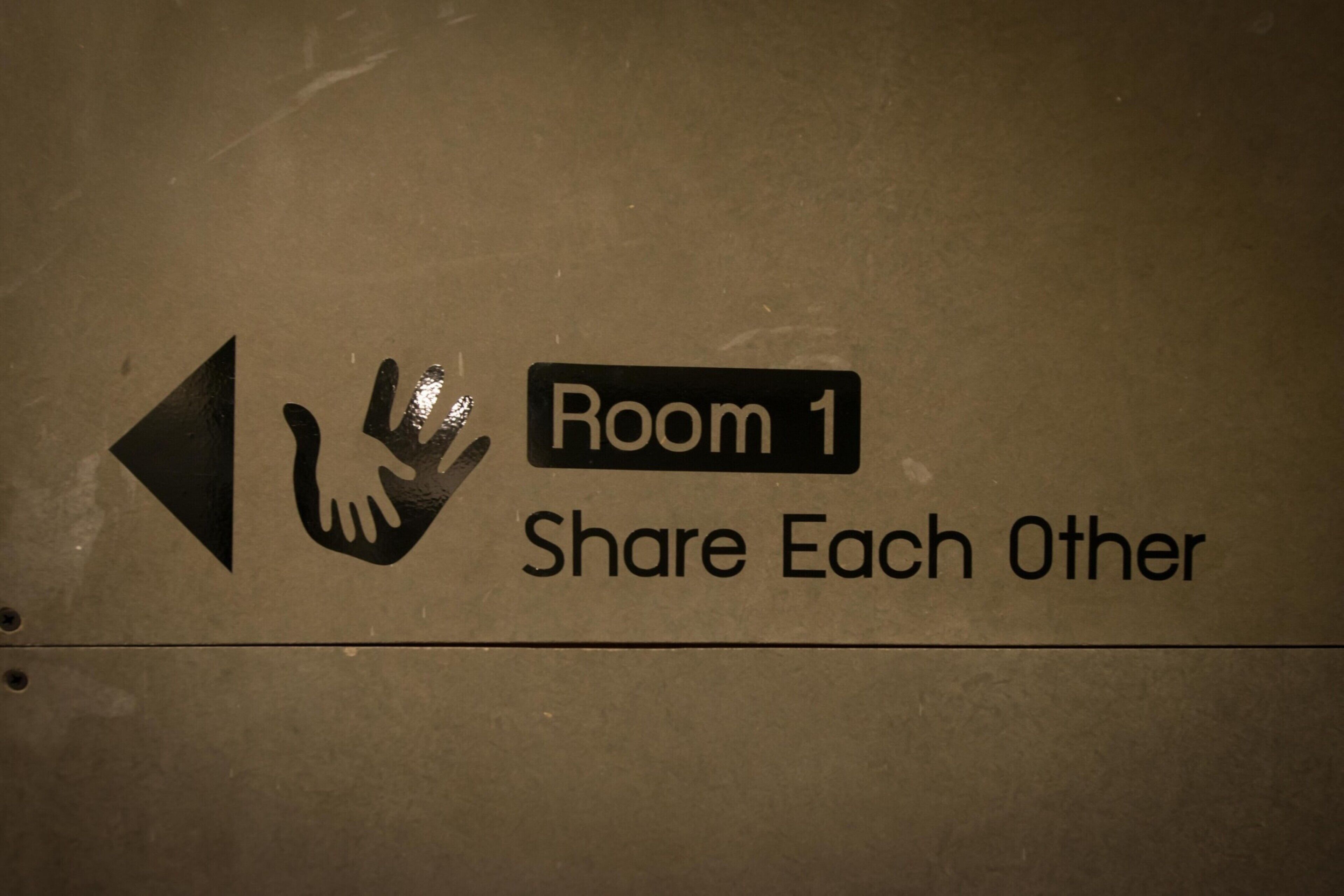 Ein Schild mit der Aufschrift 'Zimmer 1', einem Handzeichen und dem Text 'Teilt Erfahrungen'.