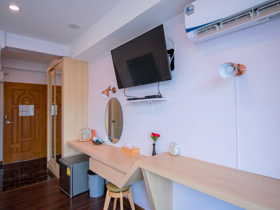 A modern hotel room with a wooden desk, a flat-screen TV mounted on the wall, a small stool, and a trash bin.