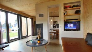 Un salon moderne avec un canapé, une table ronde sur laquelle se trouvent une bouteille et des verres, un téléviseur diffusant une chaîne d’information, et un coin cuisine visible à travers une porte ouverte.