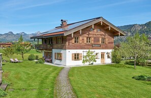 Een houten huis met twee verdiepingen, een balkon en een pad van stenen dat naar de ingang leidt, met op de achtergrond een uitzicht op bergen.