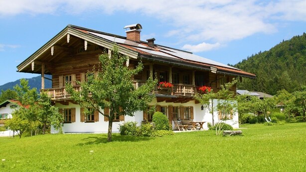 Una casa de madera tradicional con balcón, rodeada de un paisaje verde exuberante y montañas al fondo.