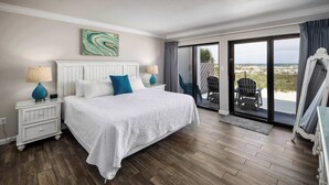 A bedroom with a large bed, two bedside tables with lamps, a view of the beach through a sliding glass door, and a balcony with chairs. - Surf Dweller 112🌊Gulf Front🏖️Free Beach Setup (Fort Walton Beach)