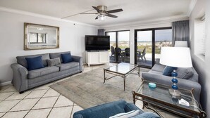 A modern living room with a grey sofa, a blue armchair, a glass coffee table, and a television. - Surf Dweller 112🌊Gulf Front🏖️Free Beach Setup (Fort Walton Beach)