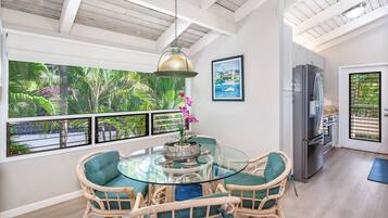 A dining area with a glass table surrounded by wicker chairs, a modern kitchen with stainless steel appliances, and large windows offering a view of lush greenery.