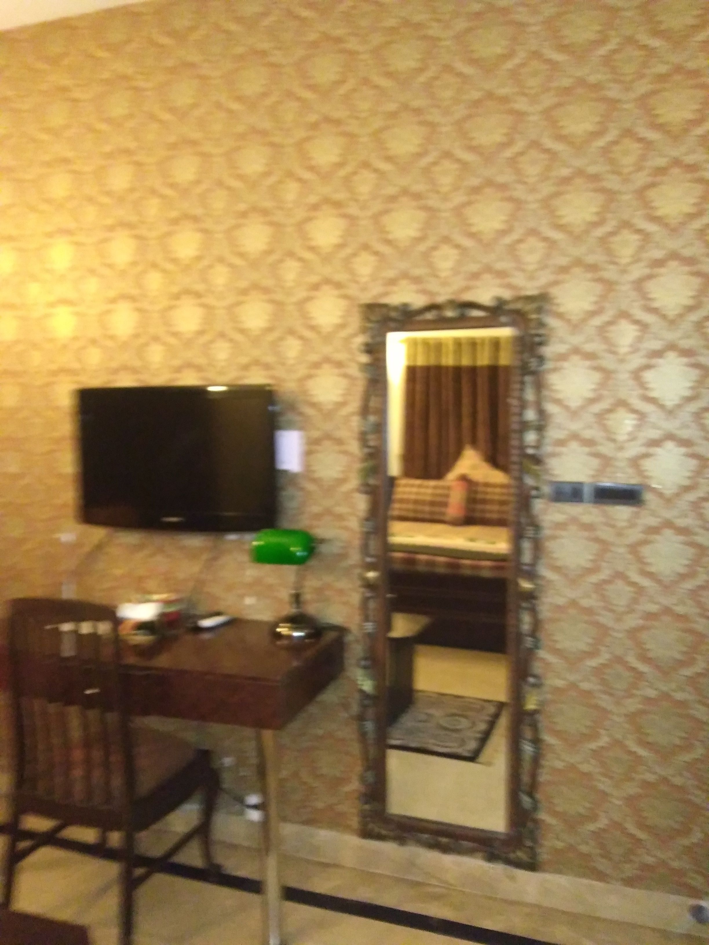A hotel room with a patterned wallpaper, a bed with a headboard, a television on a stand, a desk with a chair, and a mirror on the wall.