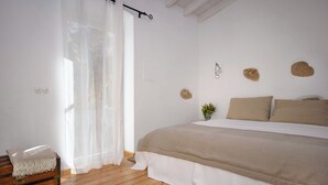 A bedroom with a wooden floor, a bed with beige bedding, a white rug, a wooden chest, and a sheer curtain.